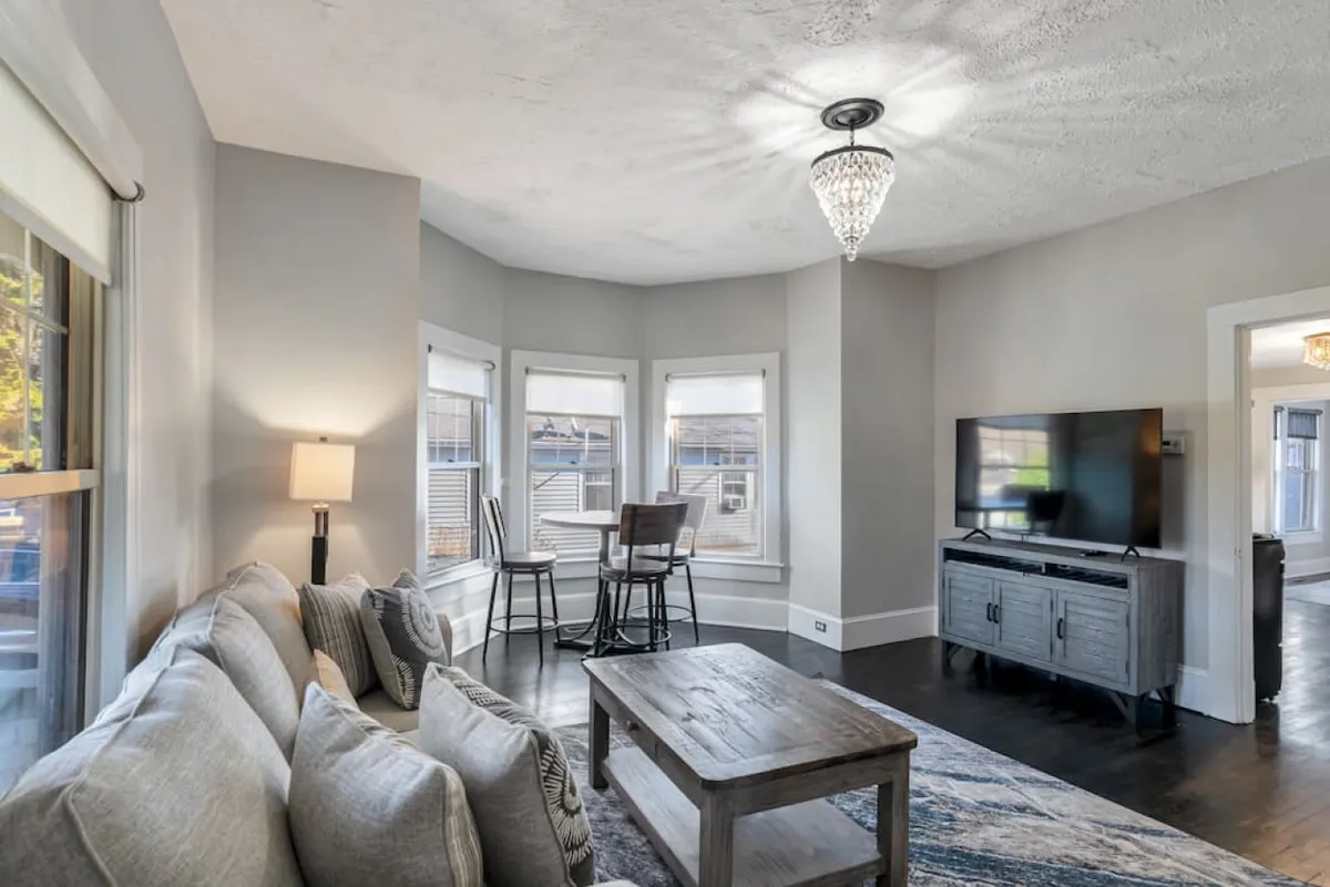 A welcoming living room featuring a plush sofa, a sturdy coffee table, a stylish chandelier overhead, and large windows that flood the space with natural light. The décor blends original architectural charm with tasteful modern updates, creating a comfortable haven for relaxing or socializing.