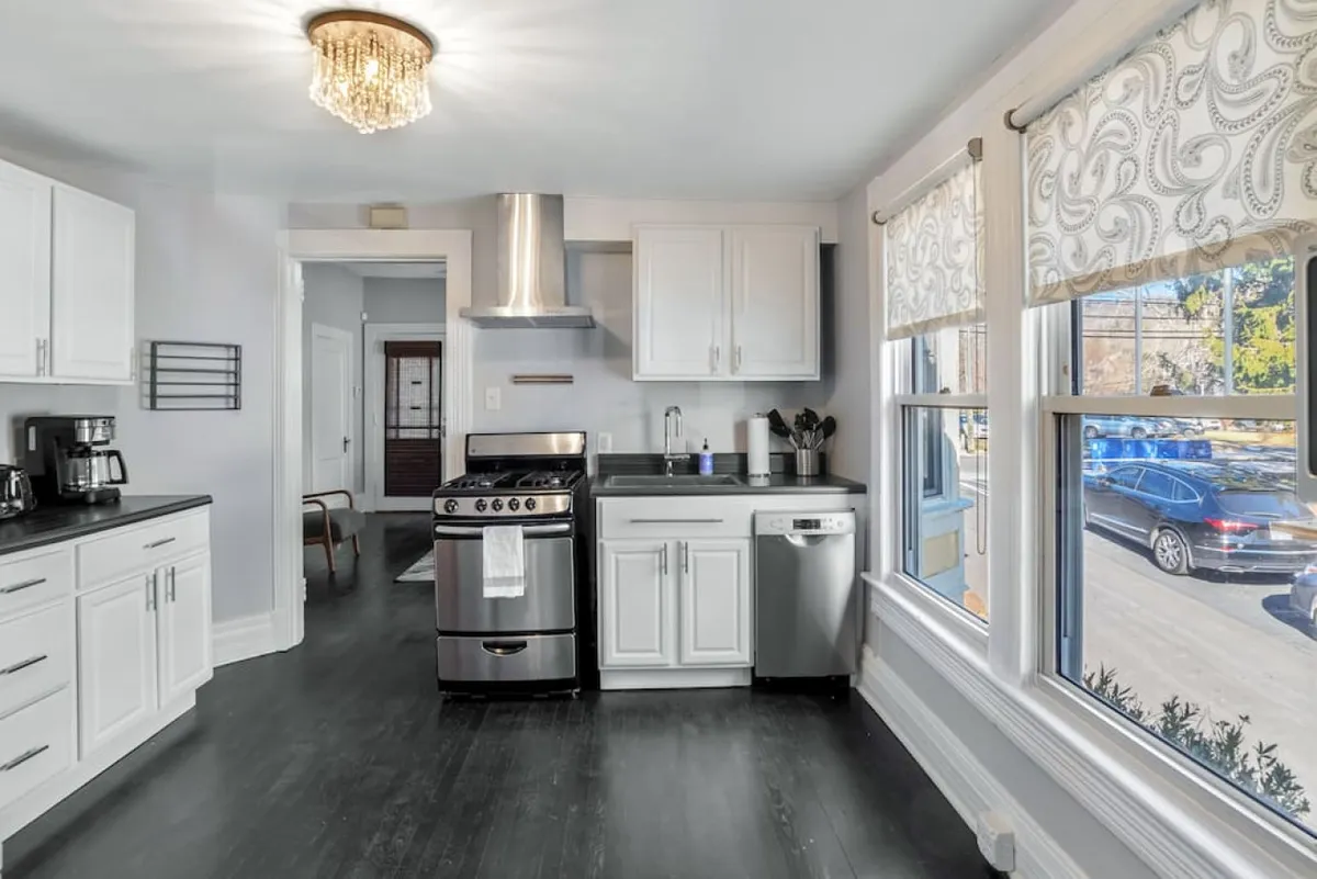 A welcoming kitchen scene featuring a stainless steel stove, crisp white cabinetry, and large windows that flood the space with light. The environment feels ideal for cooking, baking, or casual gatherings around the table.