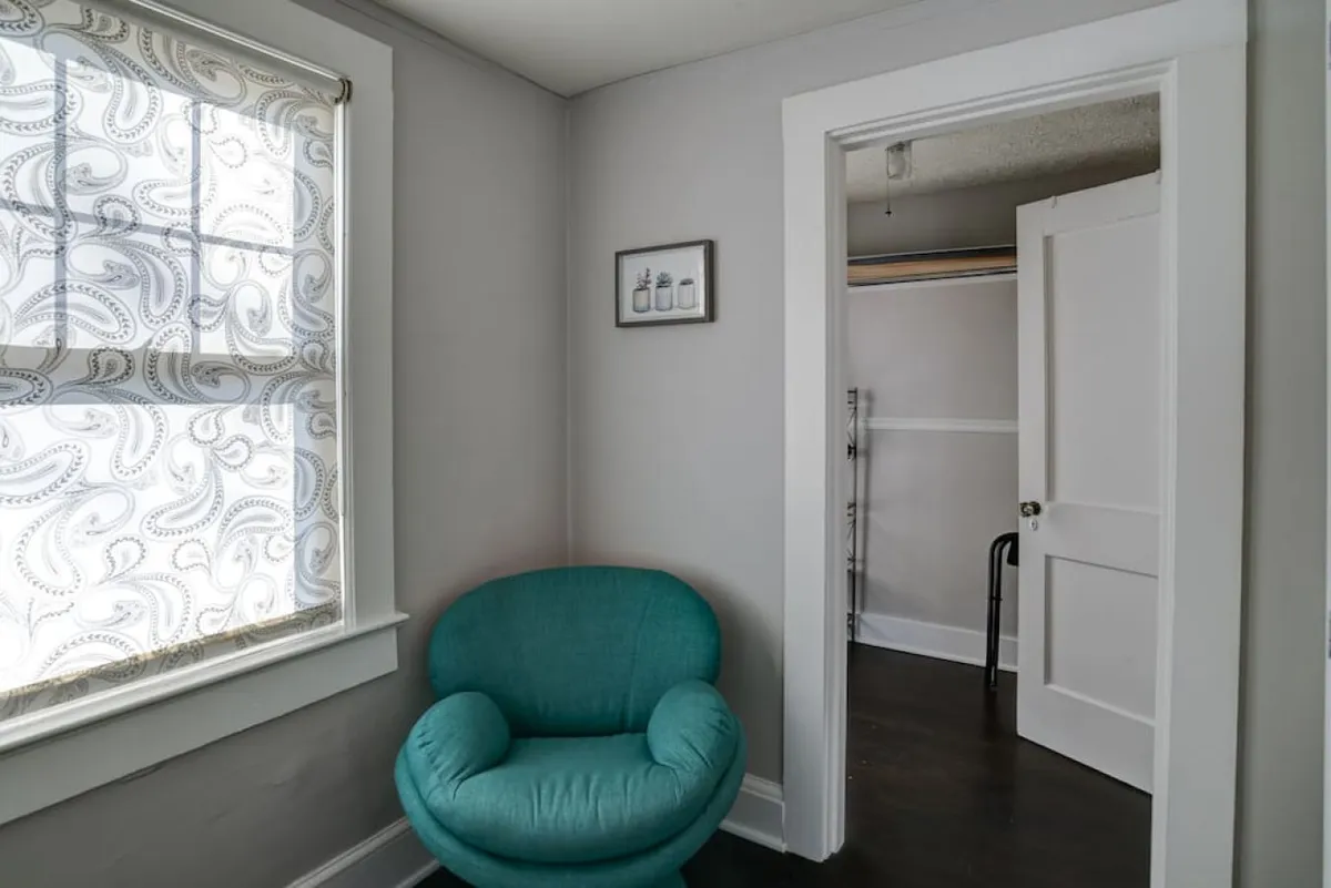 A cozy corner featuring a vibrant turquoise chair by a window adorned with patterned curtains, complemented by a piece of wall art. The intimate setting is ideal for curling up with a book or sharing a quiet conversation.
