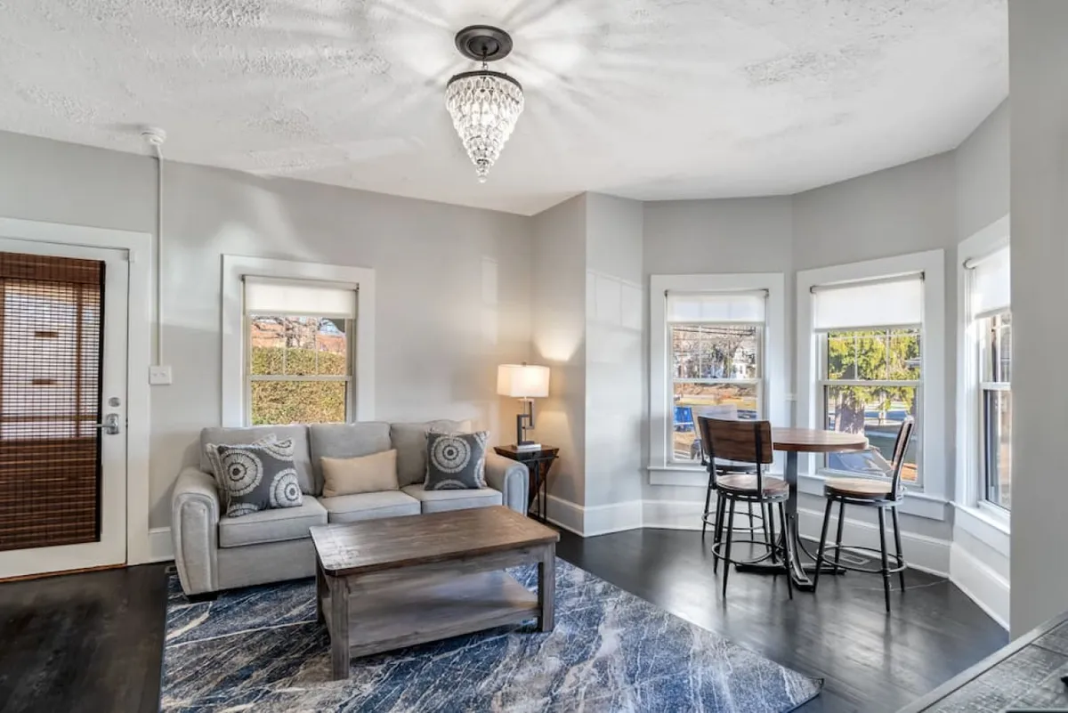 The image showcases a warm, inviting living space with a plush sofa, stylish coffee table, intimate dining area, and expansive bay windows that flood the room with natural light. The decor is tasteful and modern, set against the charm of a historic home, creating a perfect spot for relaxing, reading, or enjoying meals while taking in the neighborhood view.