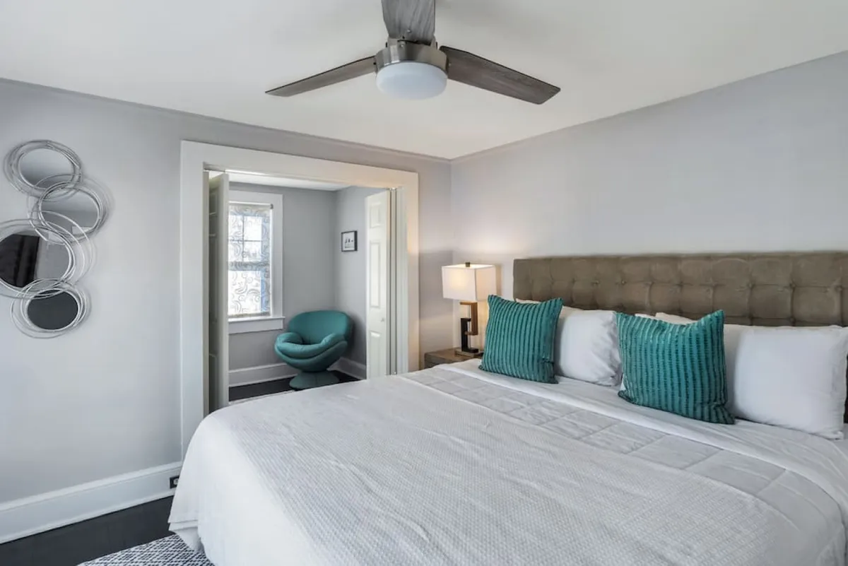 This image features a serene bedroom with a plush bed dressed in decorative pillows, a stylish lamp for soft lighting, a modern ceiling fan, a unique wall mirror, and an accent chair in the corner. The room is bathed in natural light, highlighting the inviting and restful atmosphere, and offering a cozy nook for reading or reflection.