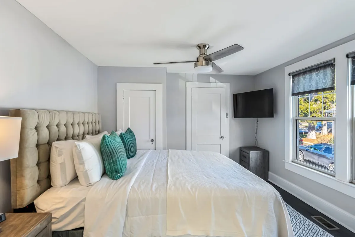 This inviting bedroom scene centers on a bed with a tufted headboard and decorative pillows, flanked by large windows that allow natural daylight to flood the space. There's a wall-mounted television, blending modern amenities with classic comforts, and the overall mood is restful and rejuvenating.