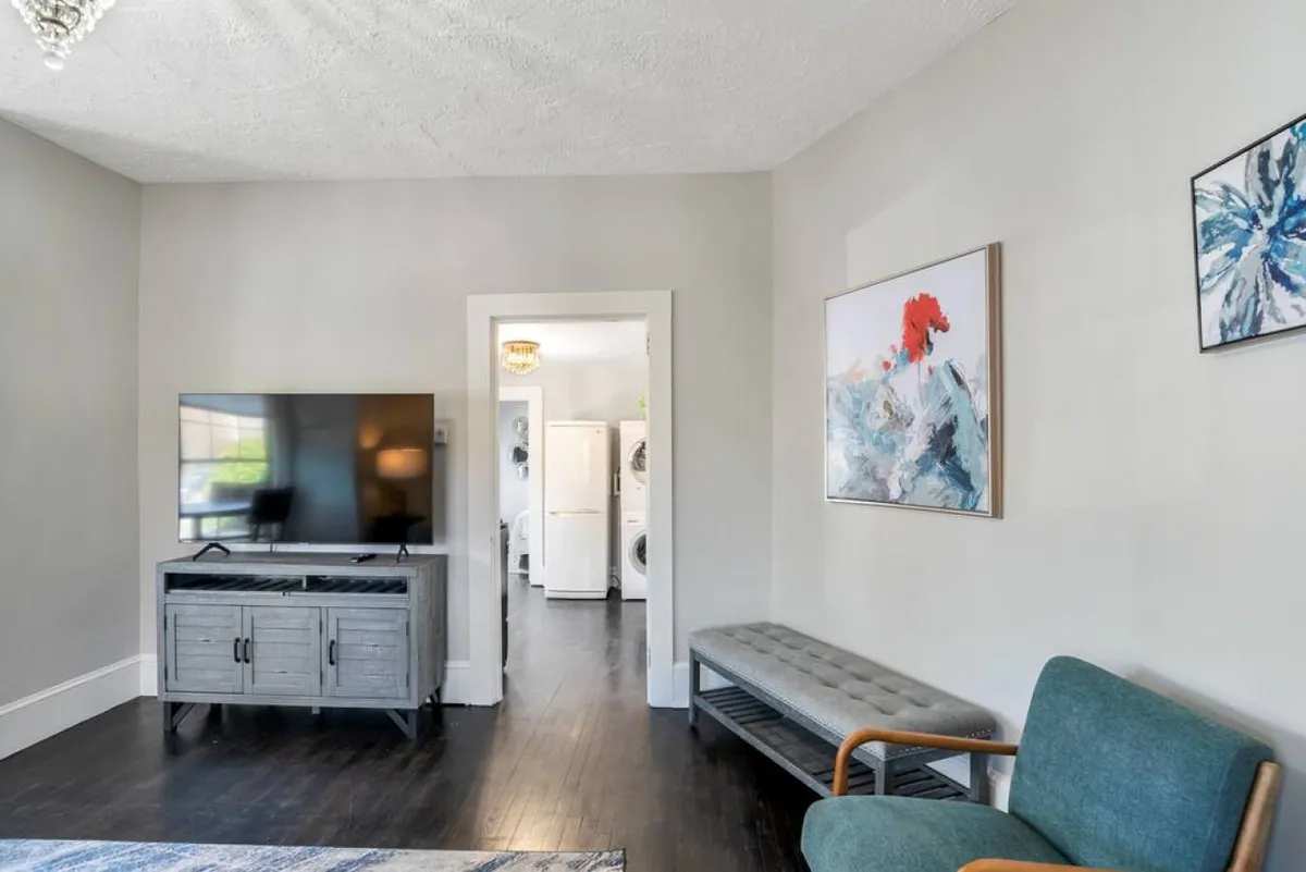 A casual, comfortable seating area anchored by a television, with attractive artwork adding color and personality, and a view toward the entrance to the laundry area. The space feels relaxed and homey, ideal for unwinding with entertainment or appreciating art.